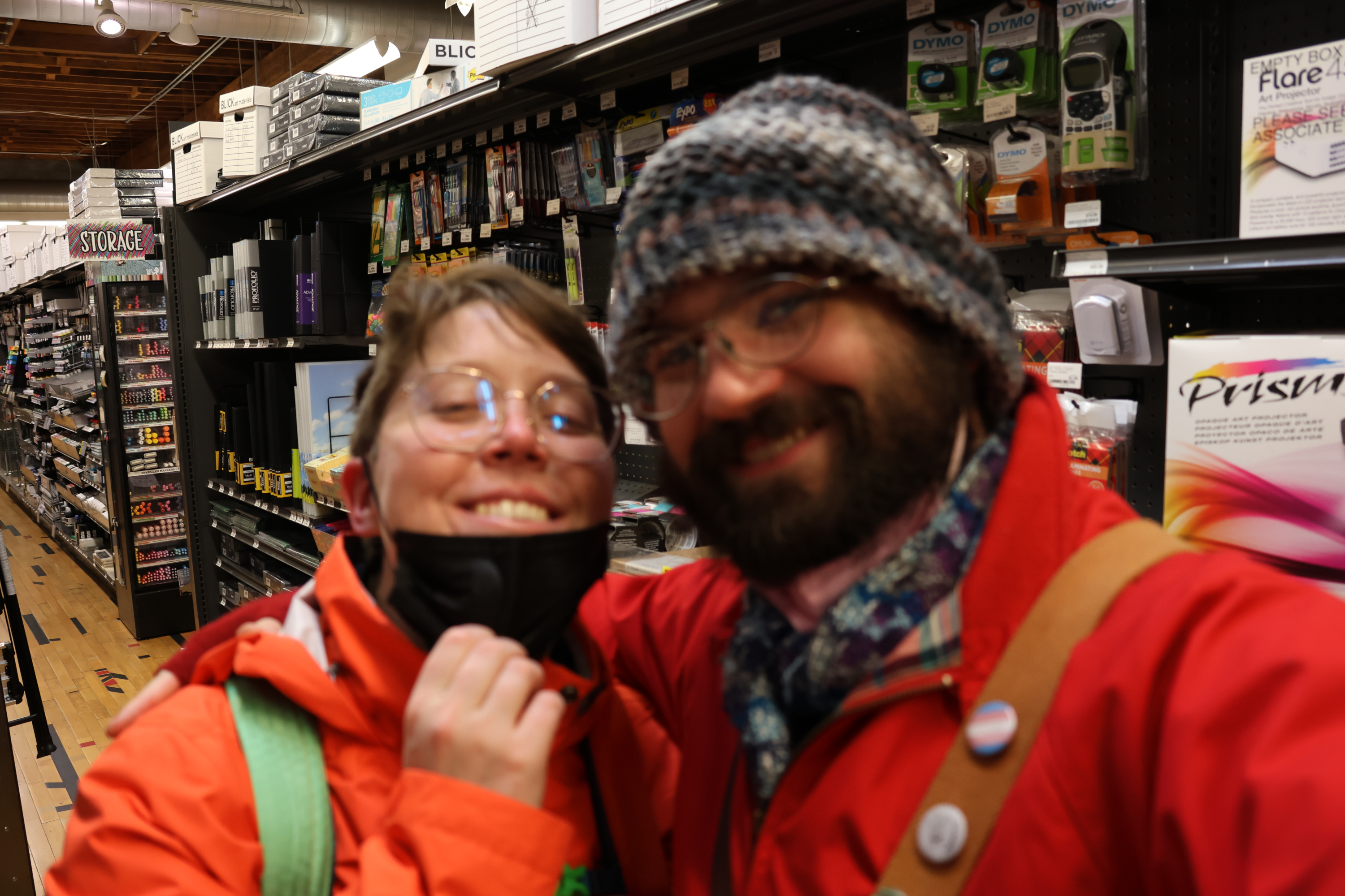 A long store aisle, shelves filled with art supplies, is in crisp focus in the background. In the foreground are Morrie and I bundled up for cold weather, pulling our masks down for a quick selfie together