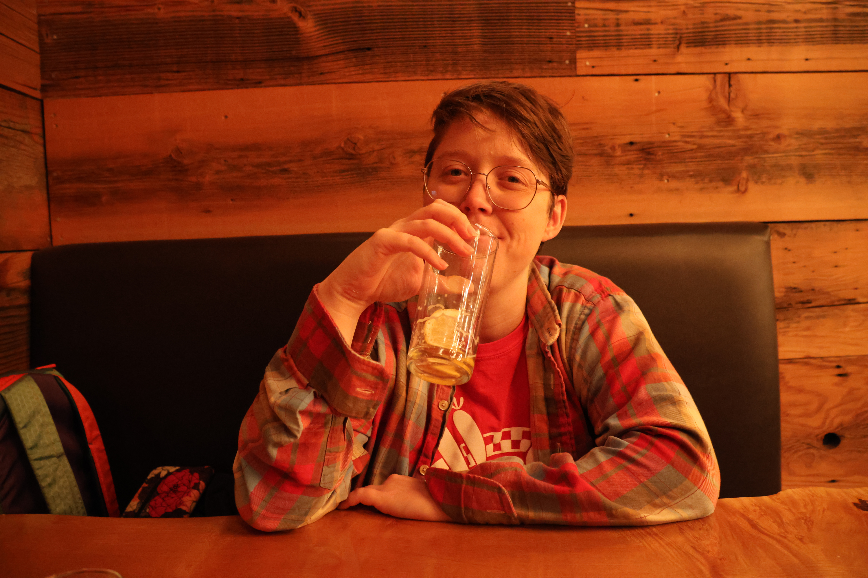 My partner Morrie sitting in a restaurant booth with rustic wood paneling behind her. She's lifting a nearly empty beer glass to her mouth, and smiling tolerantly.