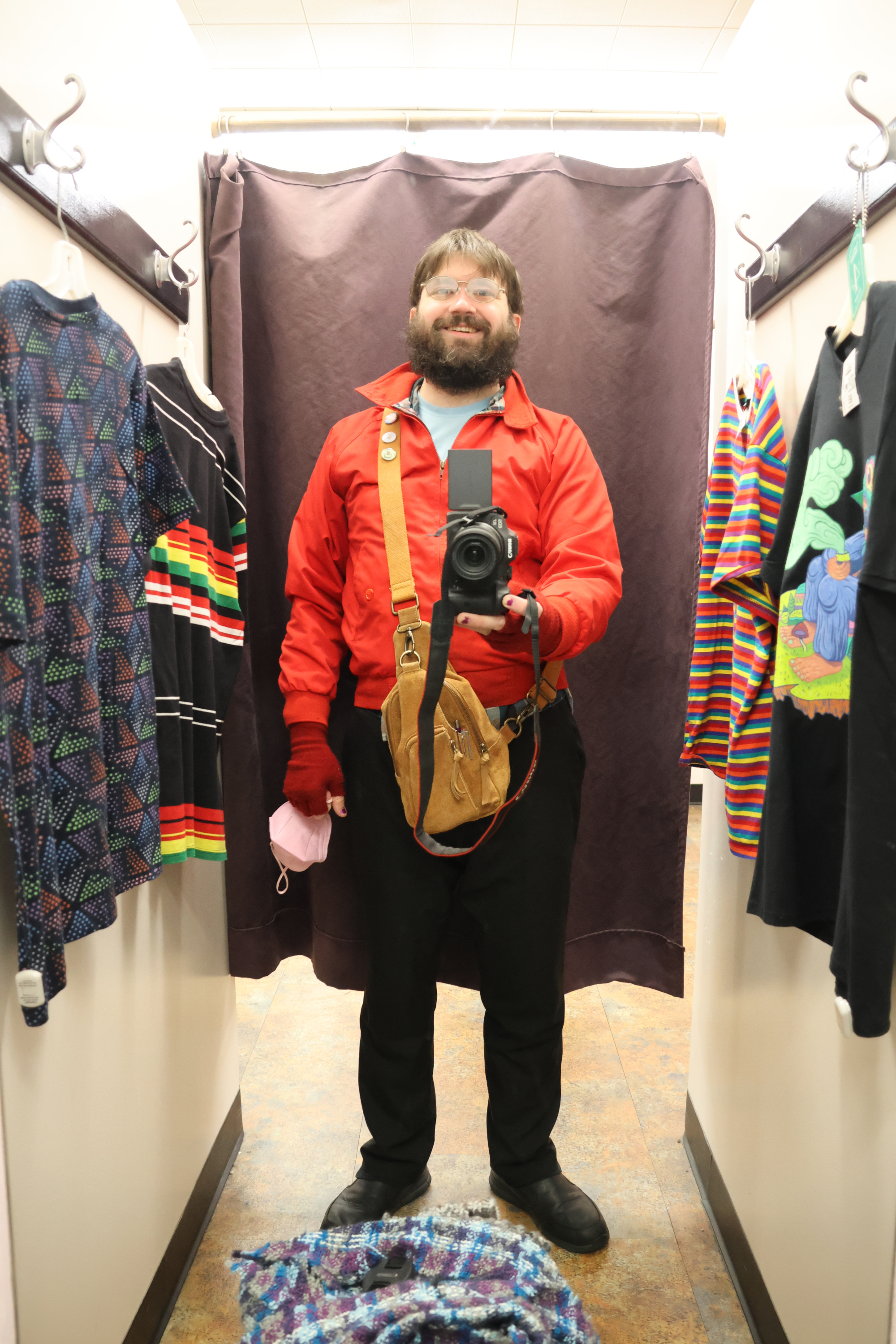 Me, bundled for cold weather, standing in a changing room. I'm pointing my camera at the mirror, and you can see four t-shirts hanging around me, ready to be tried on. From left to right: blue with colored dots arranged in triangle patterns; black with stripes of varrying thickness across the breast and waist; rainbow horizontal striped from collar to waist; a black shirt with art of a wizard smoking a pipe on it.