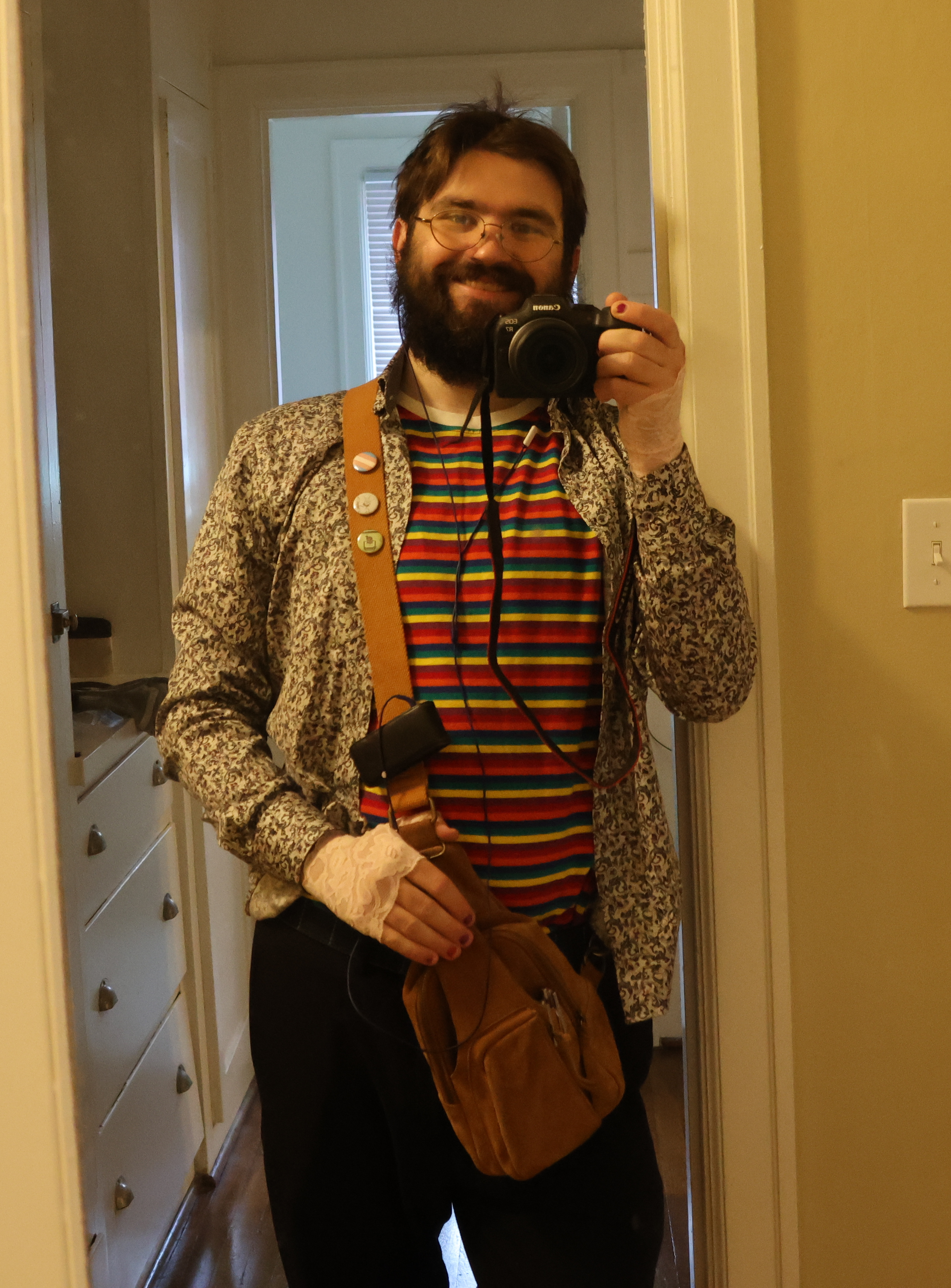 My, taking a mirror selfie. I'm wearing the rainbow striped shirt from the dressing room photo, as well as black slacks, a pattered button up worn loose and open, pink lace gloves, and a light brown satchel hanging across my torso.