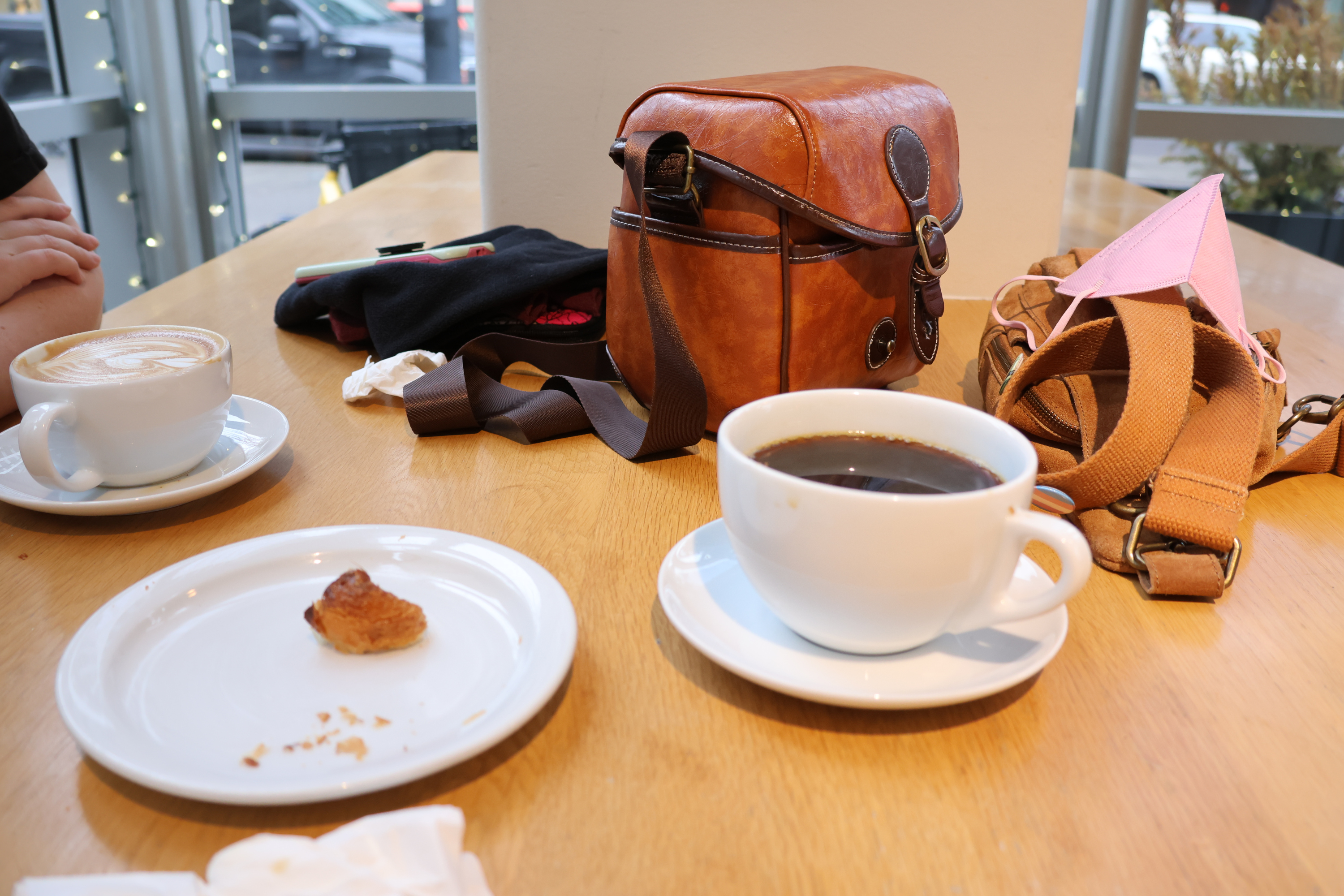 Our table at a breakfast restaurant. Our personal bags, scarfs, and Morrie's phone are on the table. A plate with the last bit of a croissant sits between us. We each have a large; mine a coffee, hers a latte.