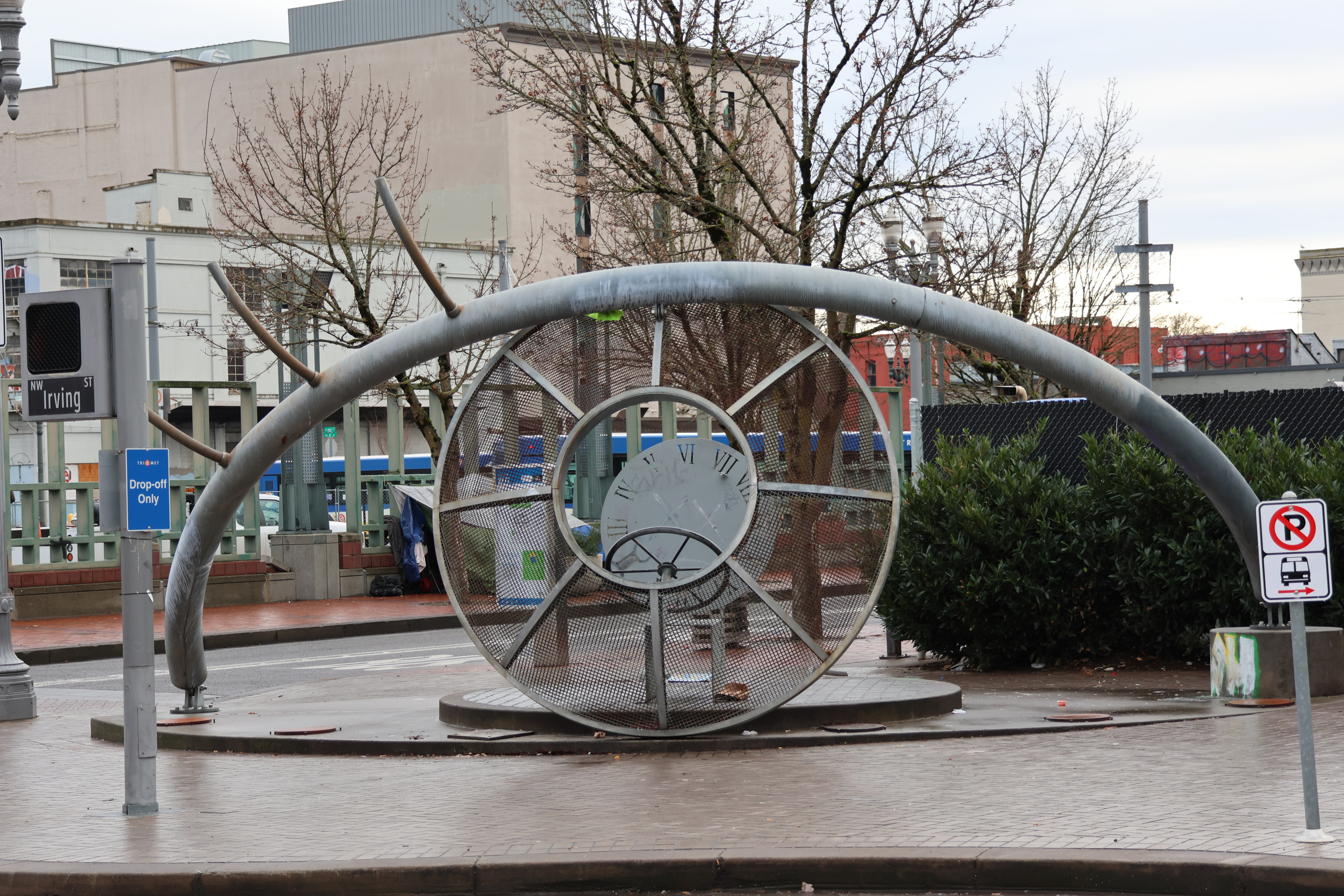 A metal sculpture of an eye, depicted only with the upper lid, 3 eyelashes, and a round metal screen for the iris. Behind the iris is a steering wheel and a seat. The back of the seat is a circle with roman numerals around its edge like a clock face.